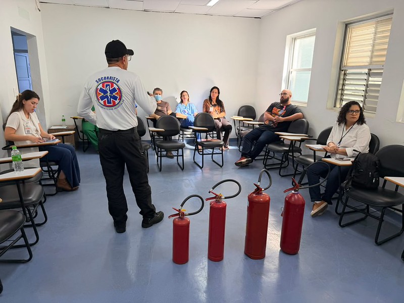 Preparar as equipes para agir de forma rápida, organizada e segura diante de situações de emergência é uma das estratégias adotadas pelo Hospital Municipal Dr. Waldemar Tebaldi, em Americana, para fortalecer sua cultura de segurança e garantir a proteção e o bem-estar de pacientes, acompanhantes, visitantes e colaboradores. Com esse objetivo, a unidade promoveu, na última semana, treinamentos sobre plano de emergência e conhecimentos de Brigada de Incêndio, reunindo profissionais de todas as áreas - assistenciais, administrativas e de apoio.
Ministrada por Jorge Machado da Silva, da MR Soluções em Saúde, a capacitação teve como foco as diretrizes da Norma Regulamentadora nº 23 (NR 23), do Ministério do Trabalho e Emprego, que estabelece as medidas de proteção contra incêndios nos ambientes de trabalho. O treinamento abordou o plano de emergência, procedimentos de prevenção, abandono de área, atuação diante de intercorrências, além de outros aspectos previstos nas normas regulamentadoras relacionadas à segurança no ambiente hospitalar. Os participantes também receberam orientações sobre as classes de incêndio e a utilização correta dos diferentes tipos de extintores.
Ao longo da atividade, os colaboradores revisaram conceitos essenciais para a identificação de situações de risco, o acionamento dos protocolos de emergência e a adoção de condutas que contribuem para minimizar riscos e garantir uma resposta rápida e coordenada em situações críticas.
"A prevenção é sempre a melhor estratégia. Quando os profissionais conhecem os procedimentos corretos e entendem seu papel dentro do plano de emergência, conseguem agir com mais segurança, organização e rapidez, reduzindo riscos e contribuindo para a proteção de todos. A segurança é uma responsabilidade compartilhada e depende da participação de cada colaborador", destacou o instrutor.
O diretor-geral do Hospital Municipal, Ruy Santos, ressalta que investir na capacitação frequente das equipes é fundamental para fortalecer a cultura de segurança, a prevenção de riscos e a melhoria contínua da qualidade dos serviços prestados. "A qualidade da assistência está diretamente relacionada à segurança e à prevenção de riscos. Por isso, investimos continuamente na capacitação das nossas equipes, para que todos estejam preparados para agir de forma rápida, organizada e segura diante de qualquer intercorrência. Esse preparo fortalece a cultura de segurança da instituição, contribui para proteger pacientes, acompanhantes, visitantes e colaboradores e reforça nosso compromisso com a melhoria contínua", afirmou.
O secretário adjunto de Saúde de Americana, Fábio Joner, reforça que a segurança é um dos pilares da qualidade na assistência em saúde. "Investir na capacitação permanente das equipes significa garantir que nossos profissionais estejam preparados para agir de forma rápida, técnica e organizada diante de qualquer situação de emergência. Esse trabalho reforça a proteção de todos, além de fortalecer a cultura de prevenção e de melhoria contínua que buscamos em toda a rede municipal de saúde", pontuou.
O Hospital Municipal Dr. Waldemar Tebaldi é administrado pelo Grupo Chavantes, em gestão compartilhada com a Secretaria Municipal de Saúde da Prefeitura de Americana.